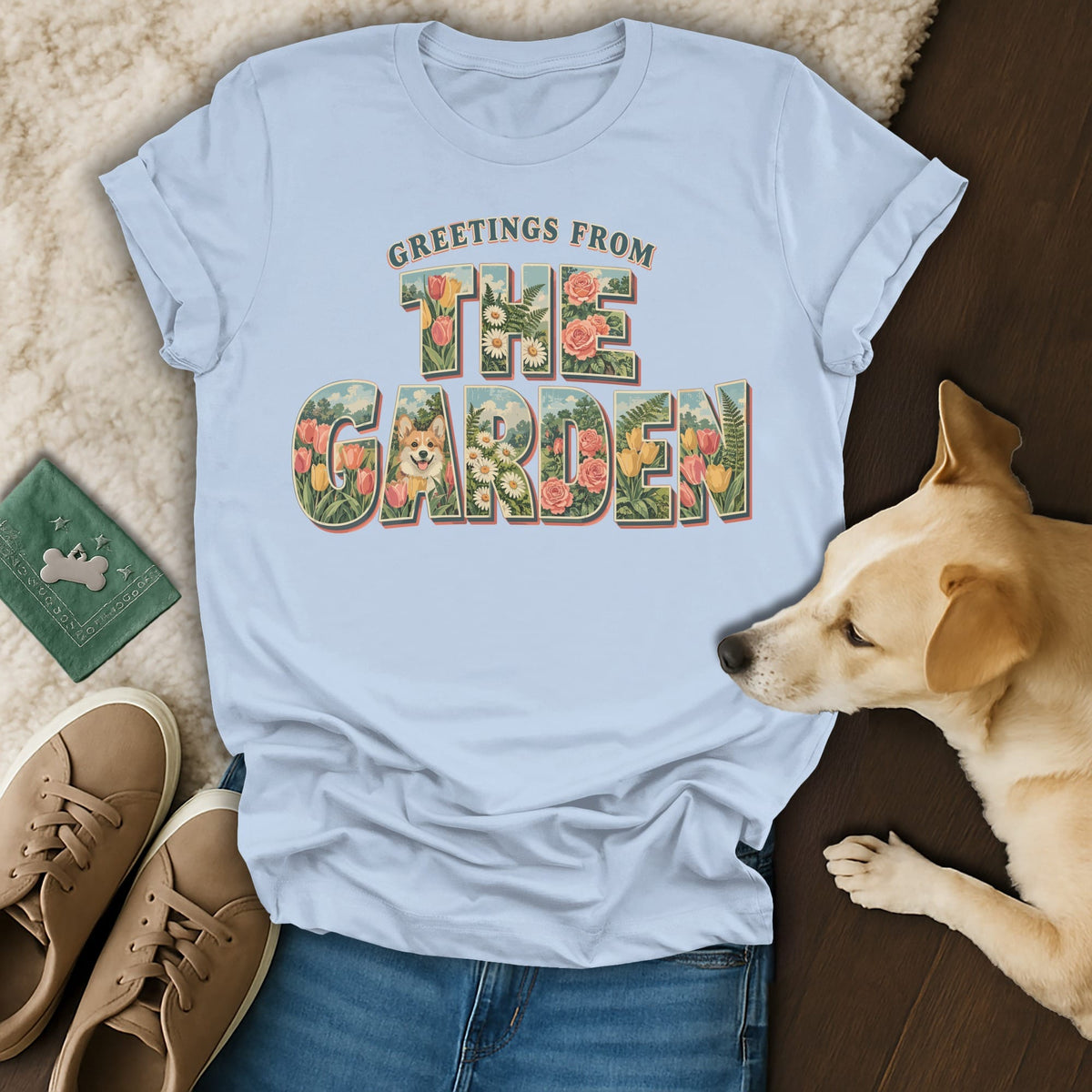 Light blue t-shirt featuring floral letters spelling Greetings From The Garden with a corgi, laid on jeans near a dog and shoes.
