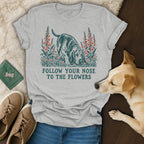 A light gray crewneck t-shirt featuring a green and pink graphic of a basshound sniffing flowers and text reading Follow Your Nose to the Flowers.