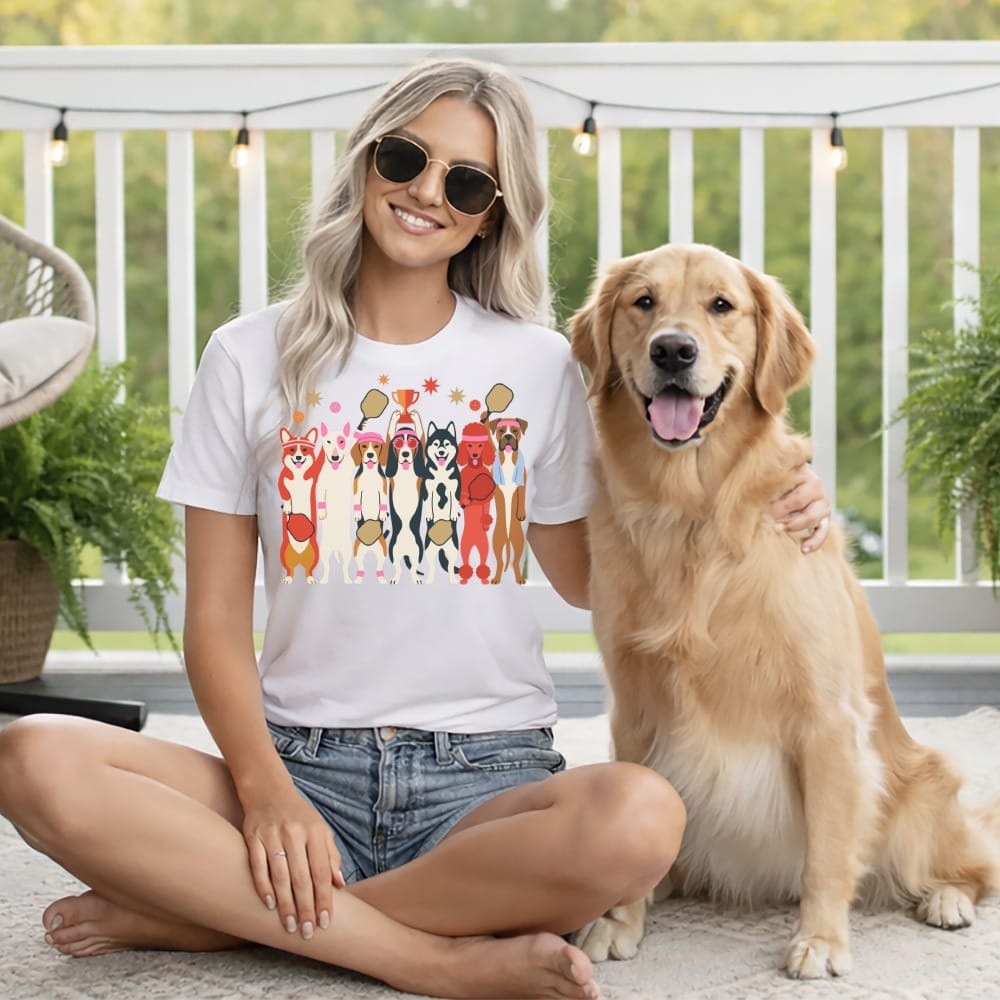 Woman in white t-shirt with cartoon dogs holding pickleballs and trophies, seated with golden retriever.
