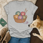 A gray t-shirt with a woven basket graphic filled with colorful yarn balls featuring paw prints.