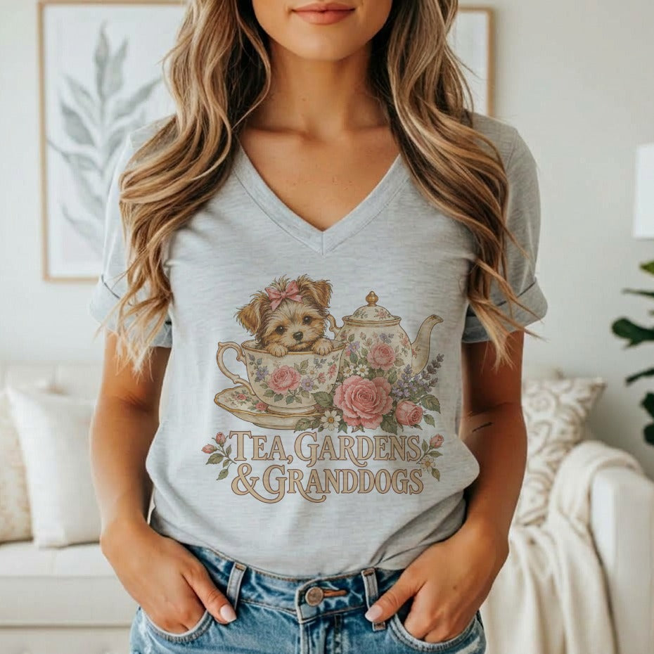 A woman wearing a gray v-neck t-shirt featuring a graphic of a puppy in a teacup, a teapot, flowers, and the text TEA, GARDENS & GRANDDOGS.