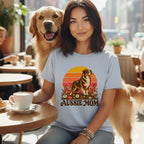 A smiling woman wearing a light blue t-shirt with an Aussie Mom graphic while seated beside a golden retriever.