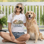 A smiling woman wearing sunglasses and a graphic t-shirt sits beside a golden retriever.