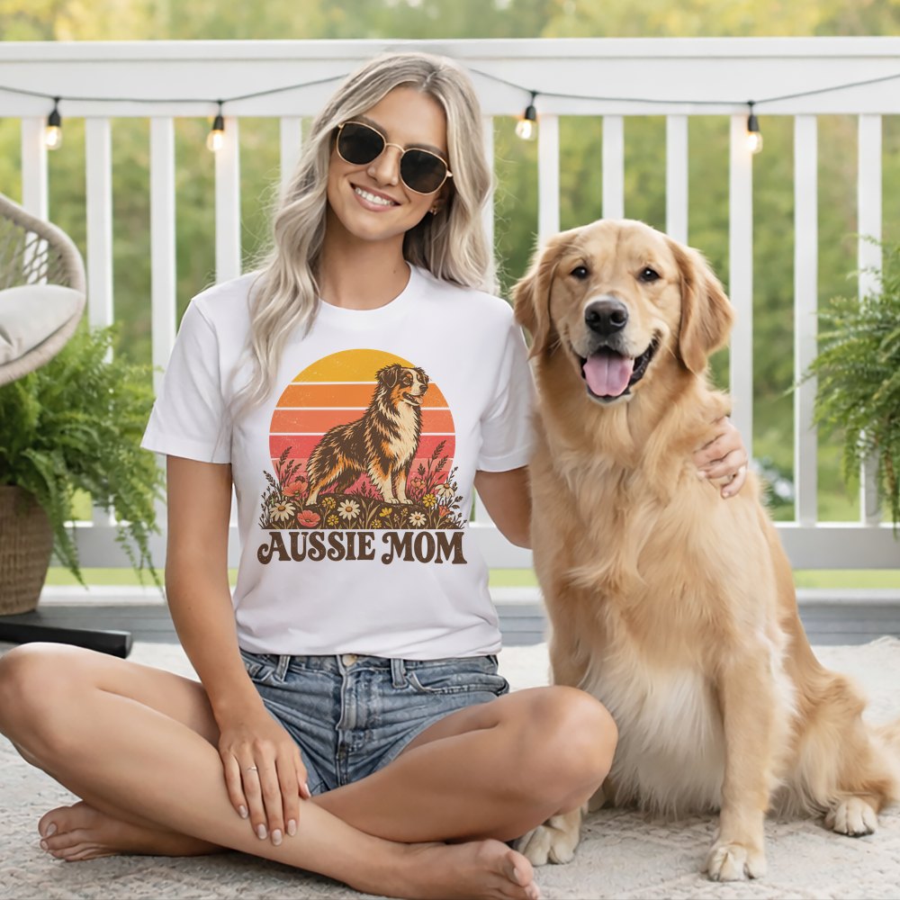 A blonde woman wearing sunglasses and a white Aussie Mom t-shirt sits with a golden retriever outdoors.