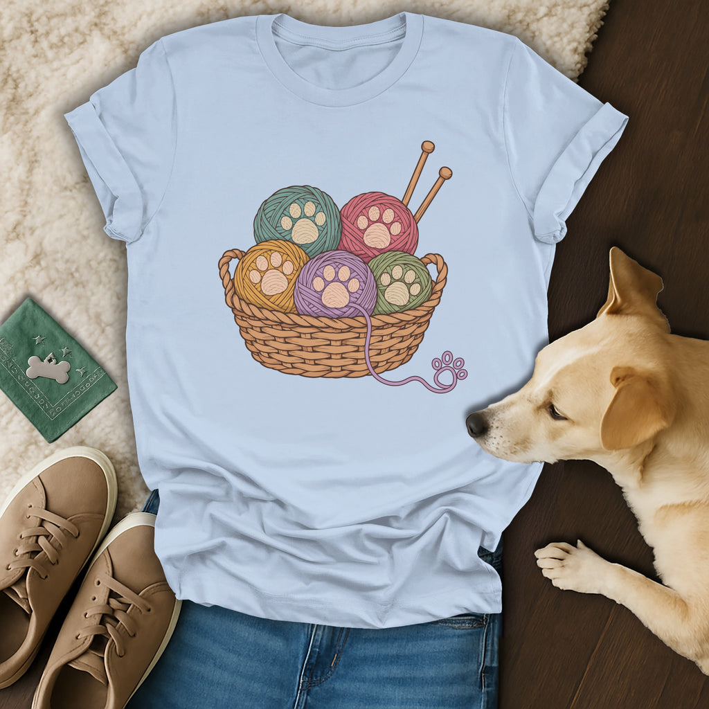 A light blue t-shirt featuring a basket of colorful yarn balls with paw print centers, laid out with a dog, shoes, and jeans.