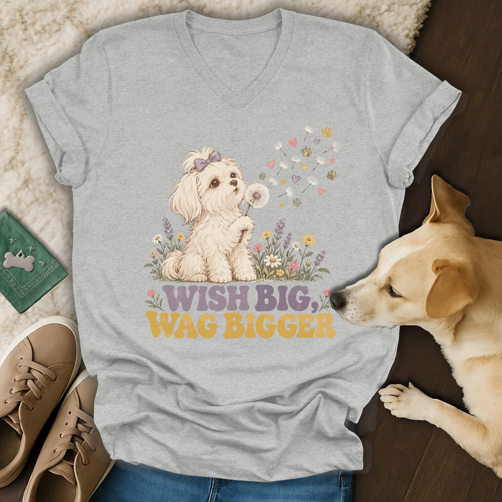 A heather gray V-neck t-shirt featuring a graphic of a fluffy white dog blowing dandelion seeds, with the text Wish Big, Wag Bigger.