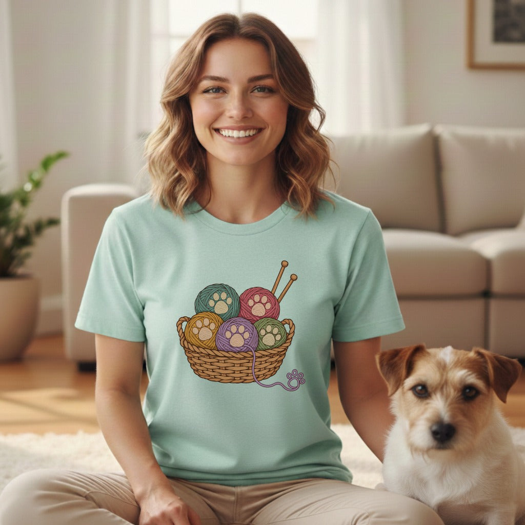 A smiling woman wearing a light blue t-shirt featuring a basket of yarn balls with paw print patterns, sitting on the floor next to a small dog in a bright living room.