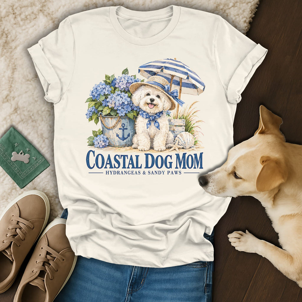 Cream t-shirt featuring a graphic of a white dog wearing a sunhat, surrounded by blue hydrangeas and an umbrella, with the text Coastal Dog Mom.