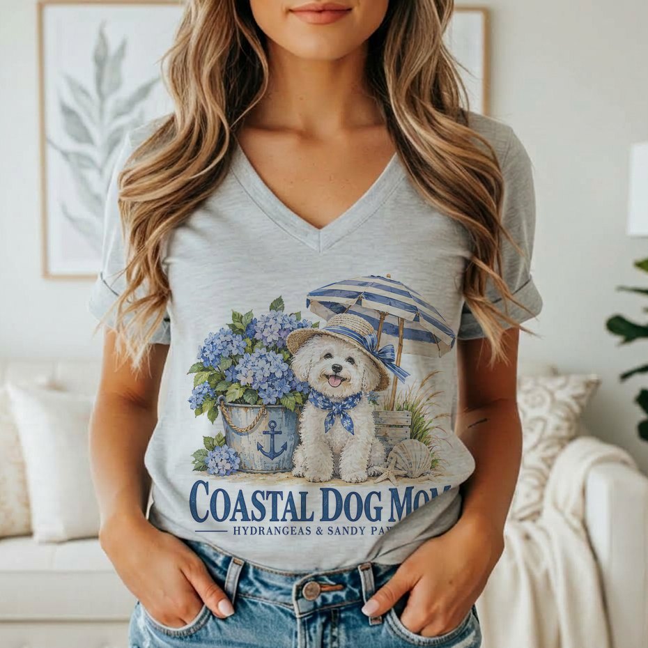 A person wearing a grey v-neck t-shirt featuring a watercolor graphic of a fluffy white dog wearing a hat and bandana, next to a bucket of blue hydrangeas and a blue and white striped umbrella, with the text "COASTAL DOG MOM / HYDRANGEAS & SANDY PAWS".