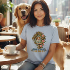 A smiling woman wearing a light blue t-shirt with a floral doghouse graphic, sitting in a cafe next to a golden retriever.