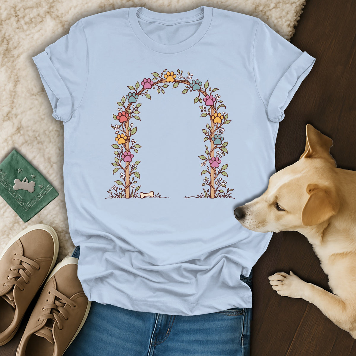 Light blue t-shirt featuring a floral arch graphic with colorful paw prints, surrounded by shoes, jeans, and a dog.