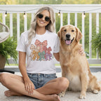 A smiling blonde woman in a white t-shirt with a colorful bow graphic sits outdoors next to a golden retriever.