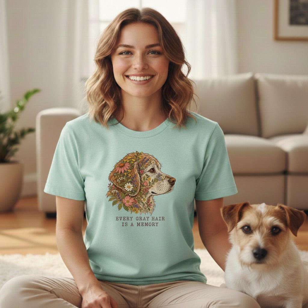 A smiling woman wears a mint green t-shirt with a floral dog graphic and text reading EVERY GRAY HAIR IS A MEMORY, sitting beside a small terrier in a living room.