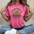 A person sitting cross-legged wears a pink t-shirt featuring a Labrador graphic, garden tools, and text.