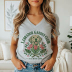 A woman wearing a heather gray V-neck t-shirt featuring a Labrador Garden Club graphic with a dog, tulips, and garden trowels.