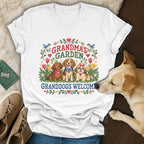White v-neck t-shirt featuring a cross-stitch graphic with three dogs, flowers, and text reading Grandmas Garden Granddogs Welcome.
