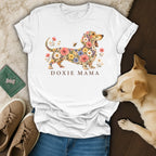 White t-shirt with a floral dachshund graphic and the text DOXIE MAMA, surrounded by accessories.