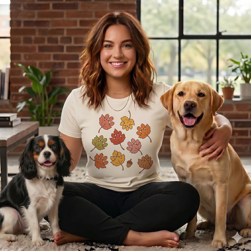 A smiling woman with a fall-leaf t-shirt sits cross-legged between a Cavalier King Charles Spaniel and a Labrador Retriever indoors.