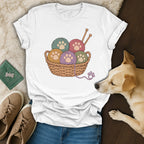 A white t-shirt featuring a graphic of colorful yarn balls with paw prints in a wicker basket, surrounded by a dog, shoes, and jeans.