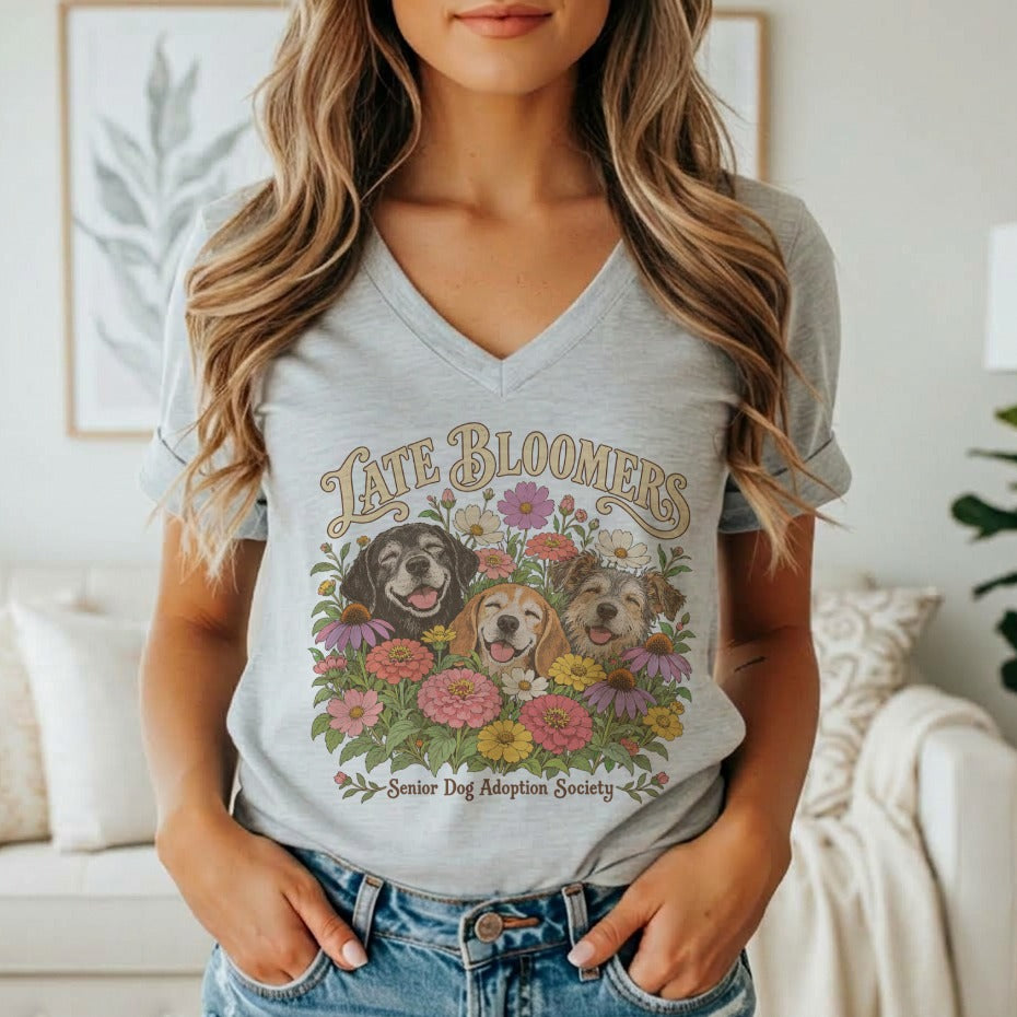 A person wearing a grey v-neck t-shirt featuring a graphic of three dogs surrounded by flowers and the text Late Bloomers Senior Dog Adoption Society.