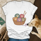 A white V-neck t-shirt featuring a basket of colorful yarn balls with paw print centers, surrounded by a dog, shoes, and jeans.
