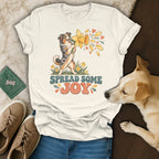 A cream-colored t-shirt featuring a retro graphic of an Australian Shepherd holding a daffodil with the text SPREAD SOME JOY, surrounded by shoes, a green wallet, and a light-colored dog on a wood and textured background.