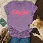 Purple t-shirt with "Snuggler" text and paw prints, next to jeans, sneakers, and a dog.