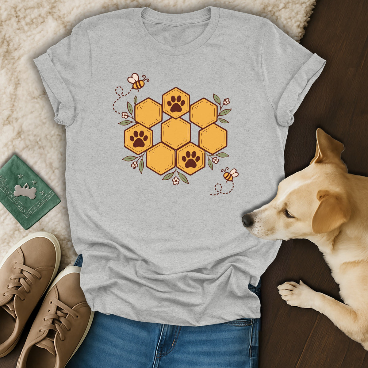 A heather gray t-shirt featuring a honeycomb graphic with paw prints and bees, surrounded by sneakers, a dog, and a green passport.