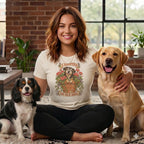 A smiling woman wearing a graphic t-shirt sits cross-legged between two dogs on the floor.