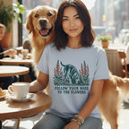 A smiling woman wears a light blue t-shirt featuring a hound graphic and the text Follow Your Nose to the Flowers while sitting with a golden retriever at a cafe.
