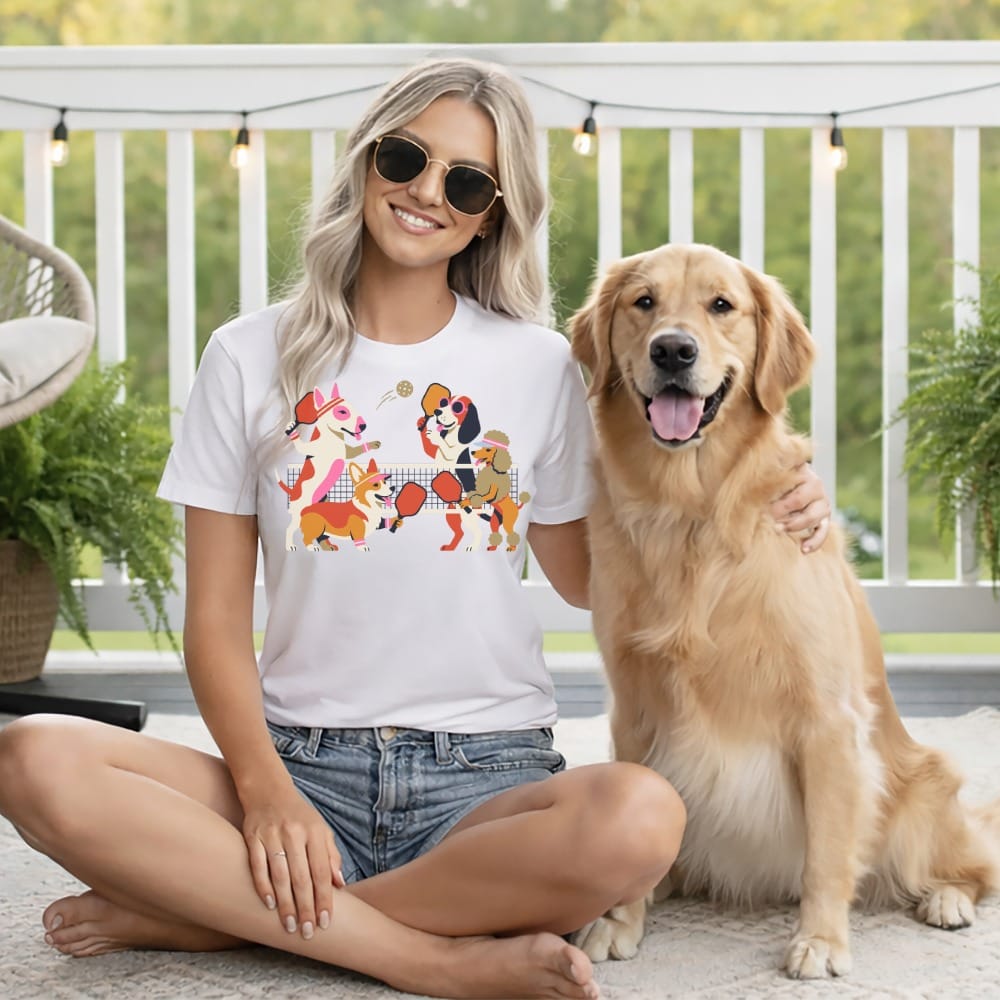Woman wearing white t-shirt with cartoon dogs playing pickleball and sitting next to a golden retriever.