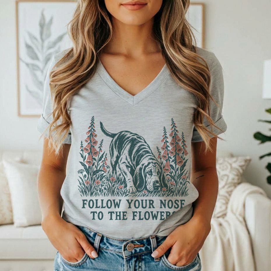A woman wearing a gray V-neck t-shirt featuring a graphic of a basshound sniffing flowers with the text FOLLOW YOUR NOSE TO THE FLOWERS.