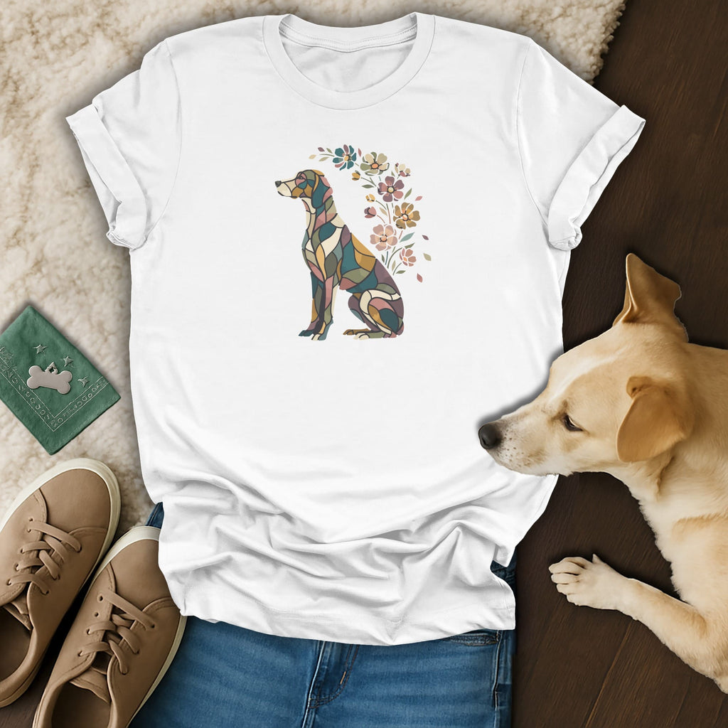 A white t-shirt featuring a mosaic-style dog graphic with flowers rests on a wooden surface next to a real dog.