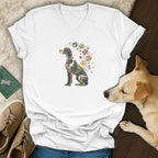 A white v-neck t-shirt features a colorful floral dog graphic, displayed with a real dog, sneakers, and accessories on a wooden surface.
