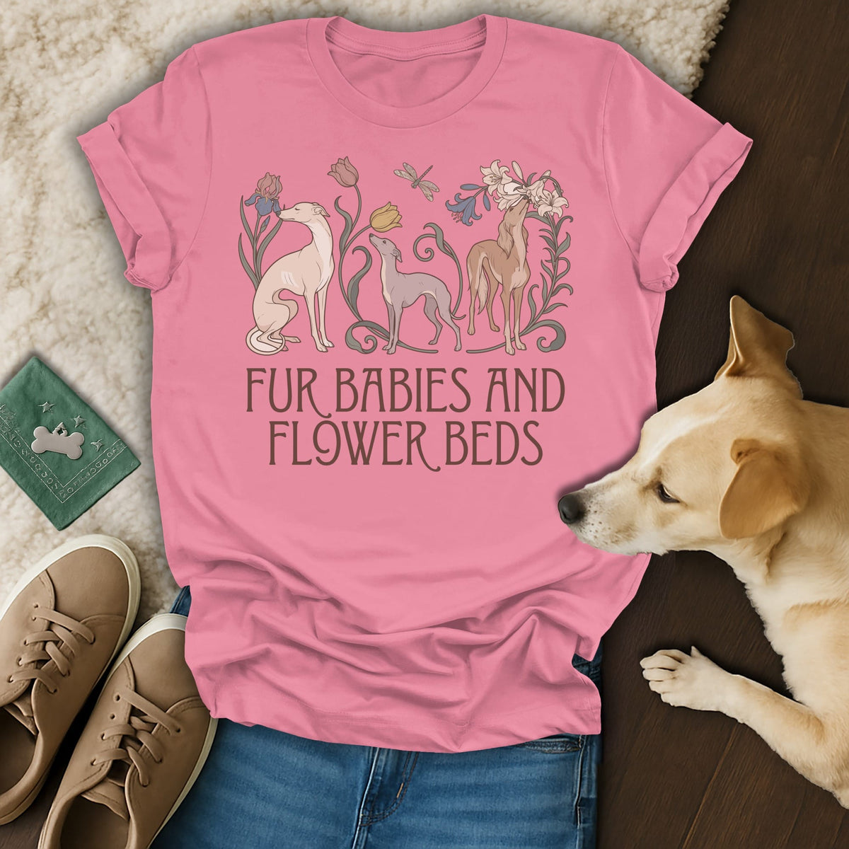 A pink t-shirt with greyhound dogs and flowers printed on the front rests on a surface next to a real dog, sneakers, and a green passport.
