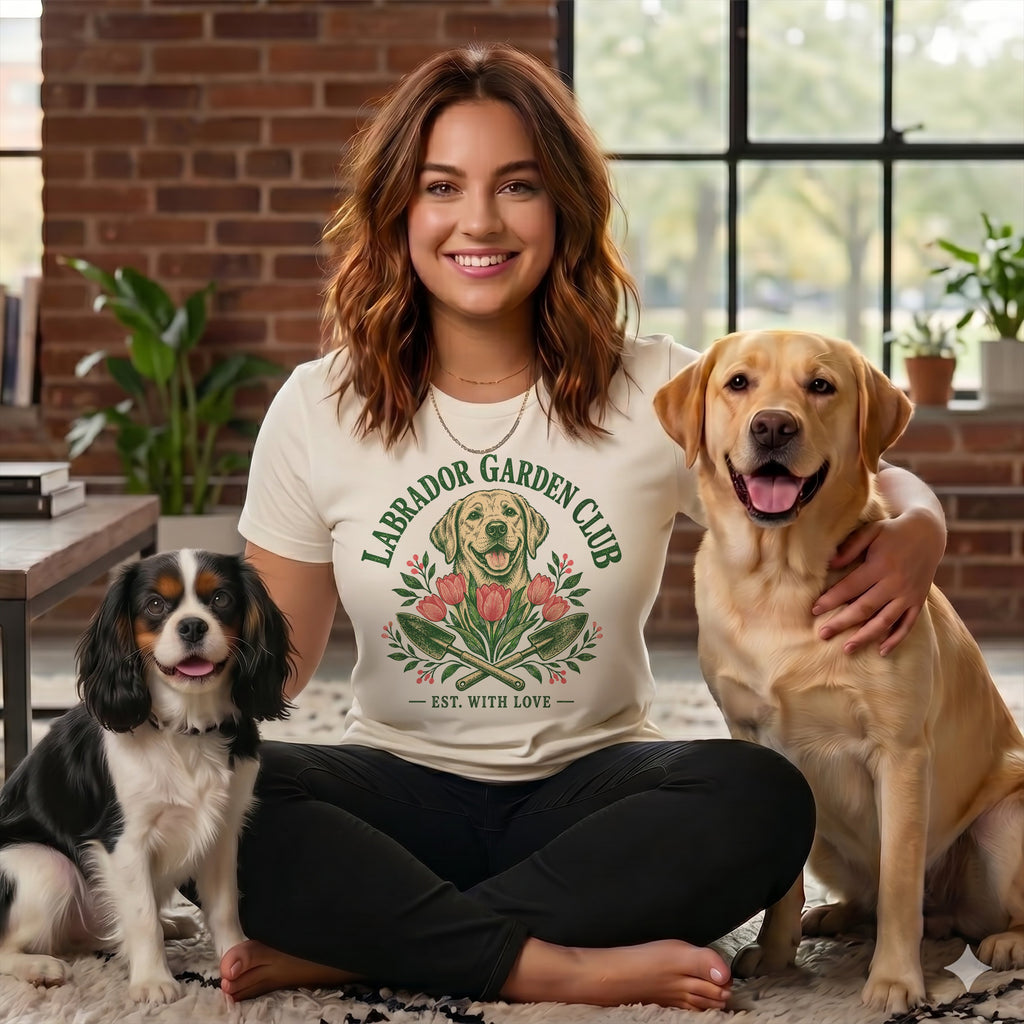 A smiling woman sits cross-legged on a rug wearing a t-shirt that says Labrador Garden Club, flanked by a King Charles Spaniel and a yellow Labrador.