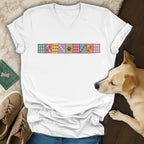 White V-neck t-shirt with a colorful patchwork design of paw prints across the chest, laid out with shoes and a dog.