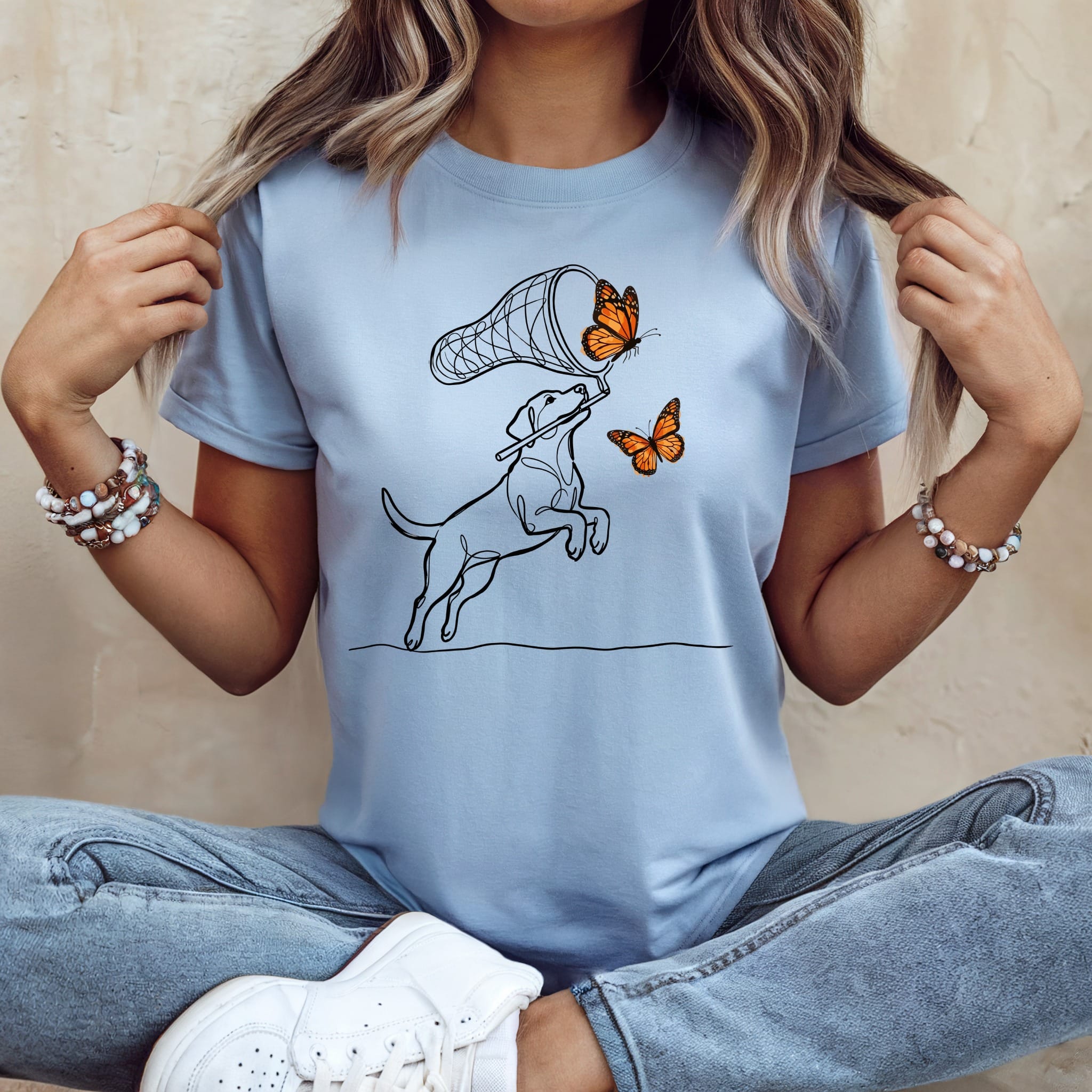 Line art of a dog chasing butterflies with a net on a light blue t-shirt.