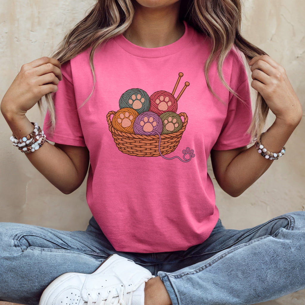 A woman wearing a pink t-shirt featuring a woven basket of colorful yarn balls with paw print designs.