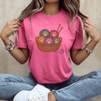 A woman wearing a pink t-shirt featuring a woven basket of colorful yarn balls with paw print designs.
