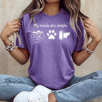 Purple t-shirt with white text "My needs are simple" and graphics of a camera, paw print, and film roll.