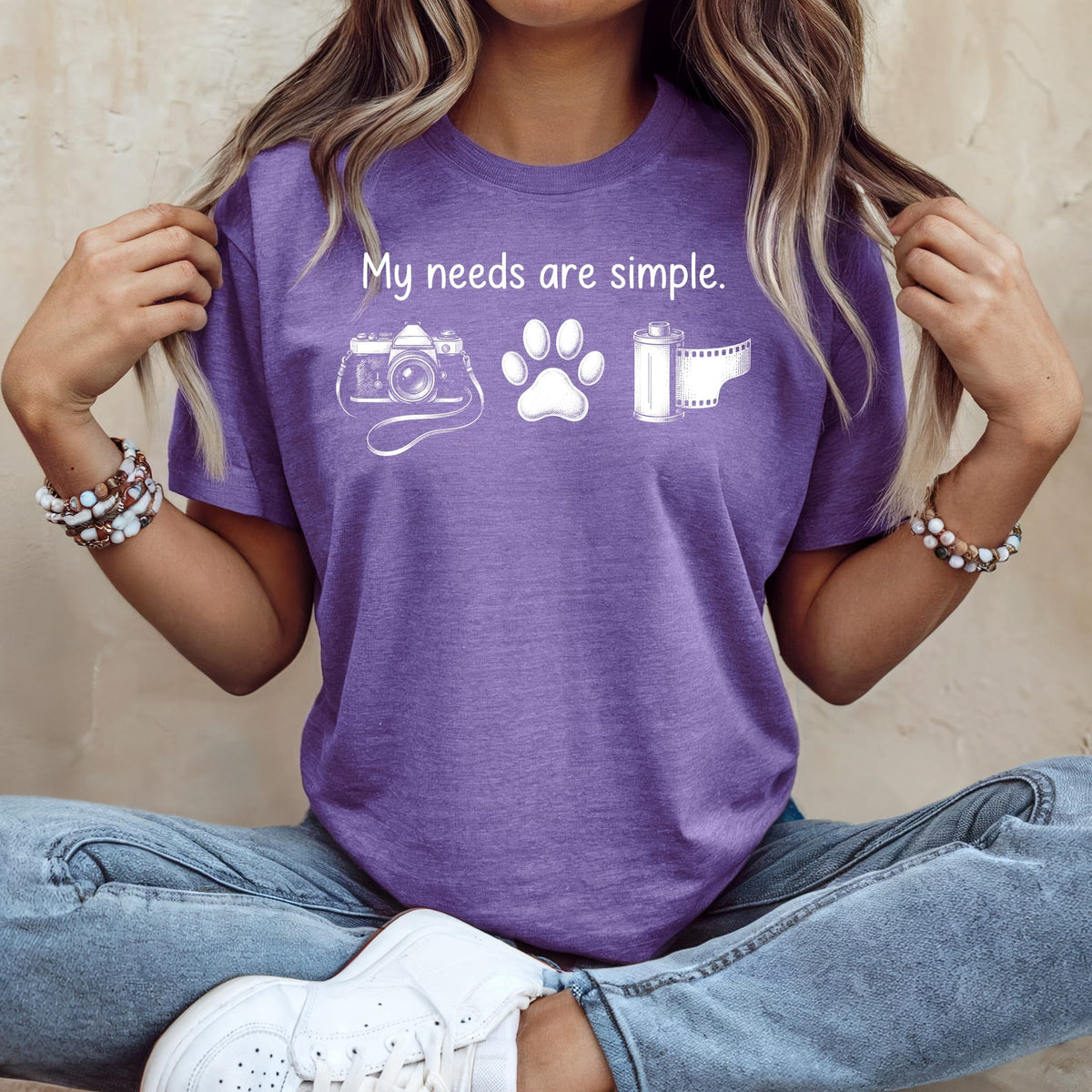 Purple t-shirt with white text "My needs are simple" and graphics of a camera, paw print, and film roll.