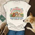 A white t-shirt featuring a cross-stitch design with dogs, flowers, and the text Grandma's Garden Granddogs Welcome.