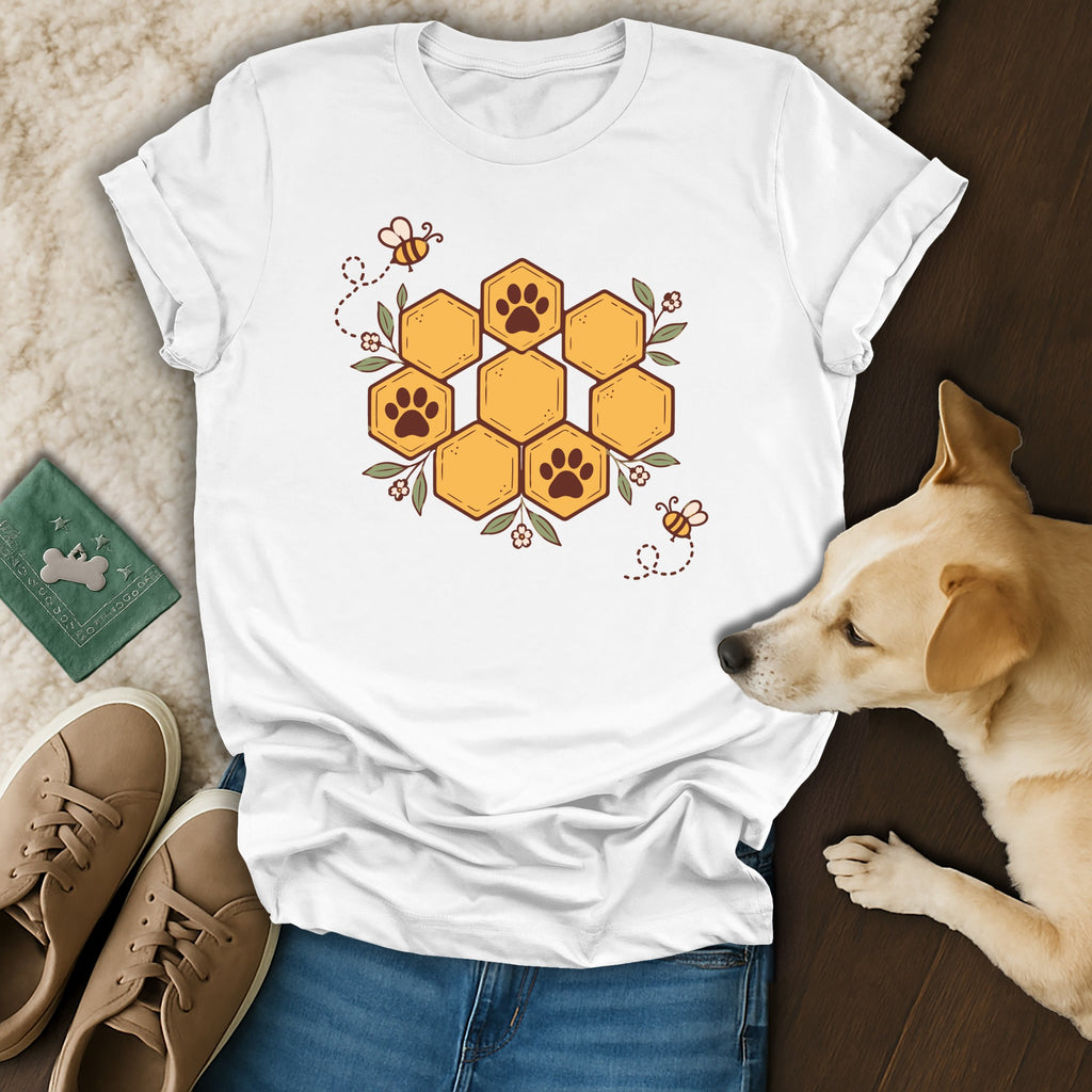 A white t-shirt featuring a honeycomb graphic with paw prints, bees, and flowers lies flat alongside blue jeans, brown sneakers, and a small dog resting on a wooden surface.