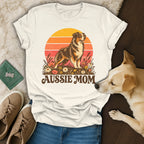 A cream-colored t-shirt with an Australian Shepherd graphic, text reading AUSSIE MOM, surrounded by casual accessories and a real dog.