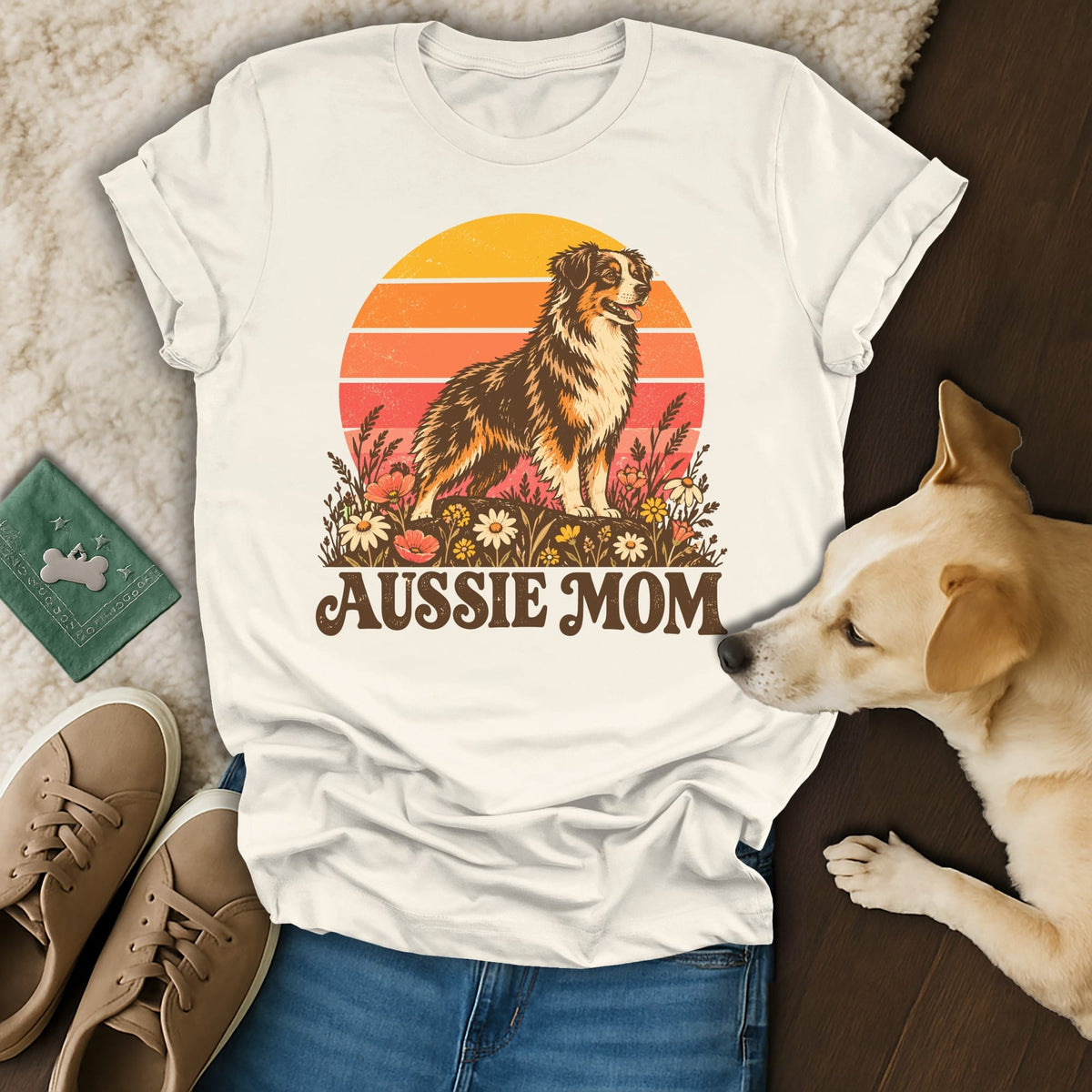 A cream-colored t-shirt with an Australian Shepherd graphic, text reading AUSSIE MOM, surrounded by casual accessories and a real dog.