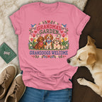 A pink t-shirt featuring a cross-stitch design of three dogs, flowers, and the text Grandma's Garden Granddogs Welcome.