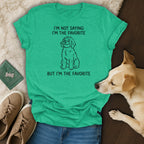Green t-shirt with black text "I'M NOT SAYING I'M THE FAVORITE BUT I'M THE FAVORITE" and dog illustration.
