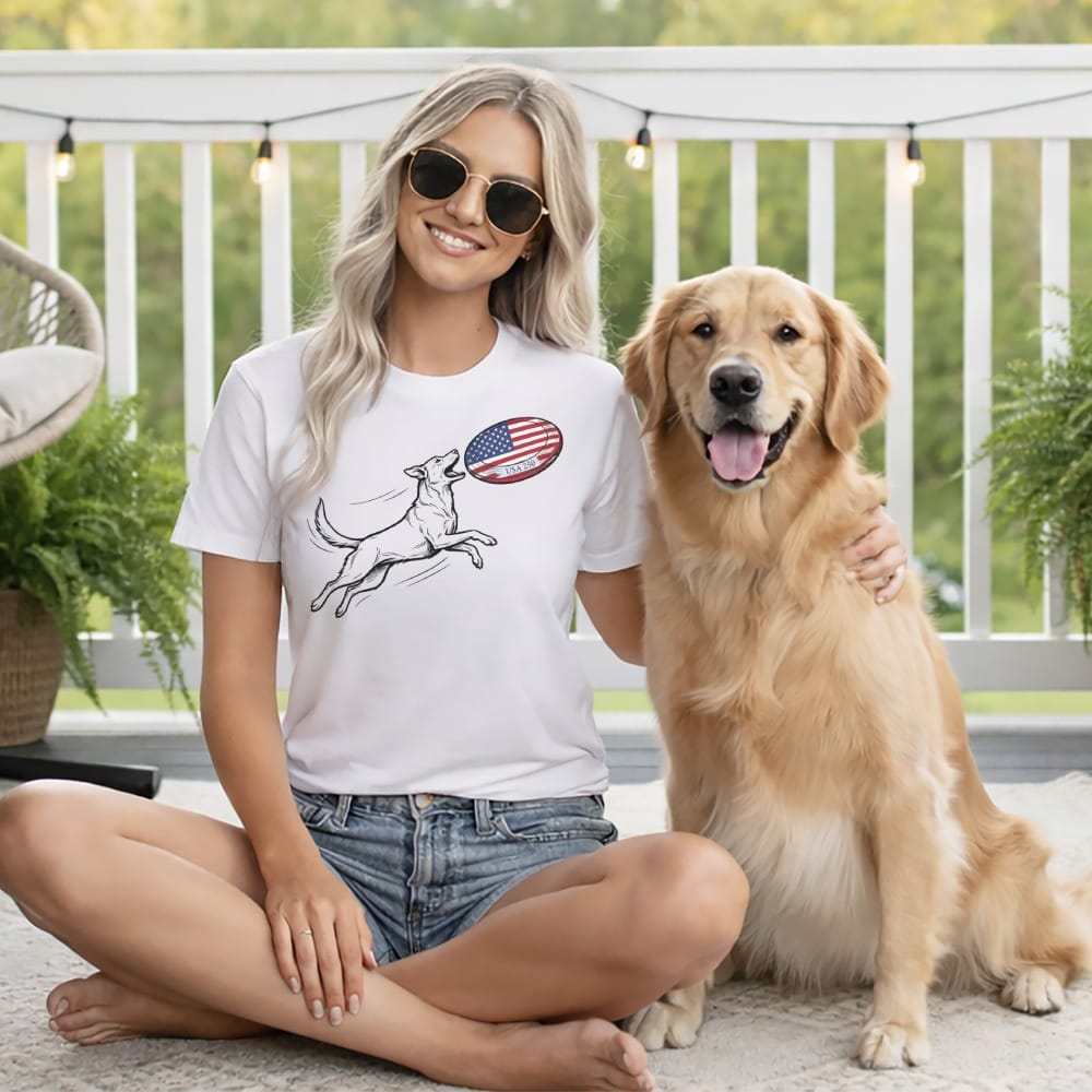 Woman wearing a white t-shirt with a dog and American flag frisbee graphic, sitting with Golden Retriever.