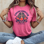 A person wearing a pink t-shirt with a Cavalier Garden Club graphic featuring a dog, roses, and gardening trowels.